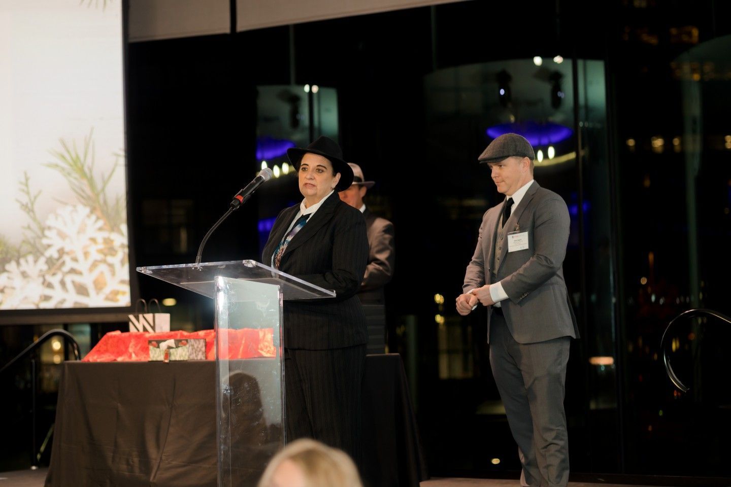 Woman speaking at a podium, man in hat stands beside her at an event. Dark setting, city lights visible.