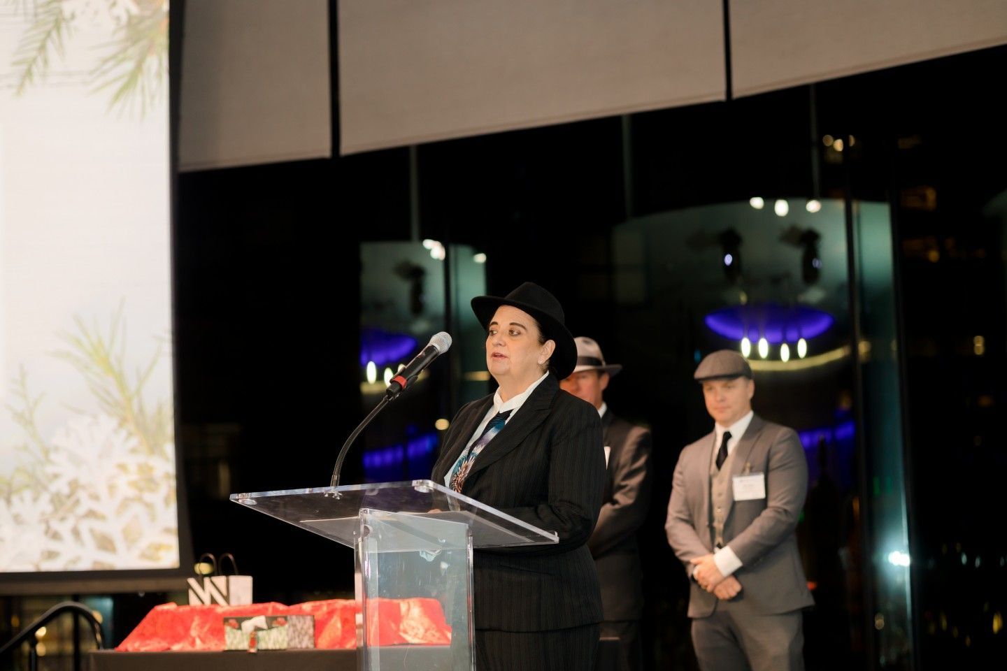 Woman in a suit and hat speaking at a podium; two men in suits stand behind her.