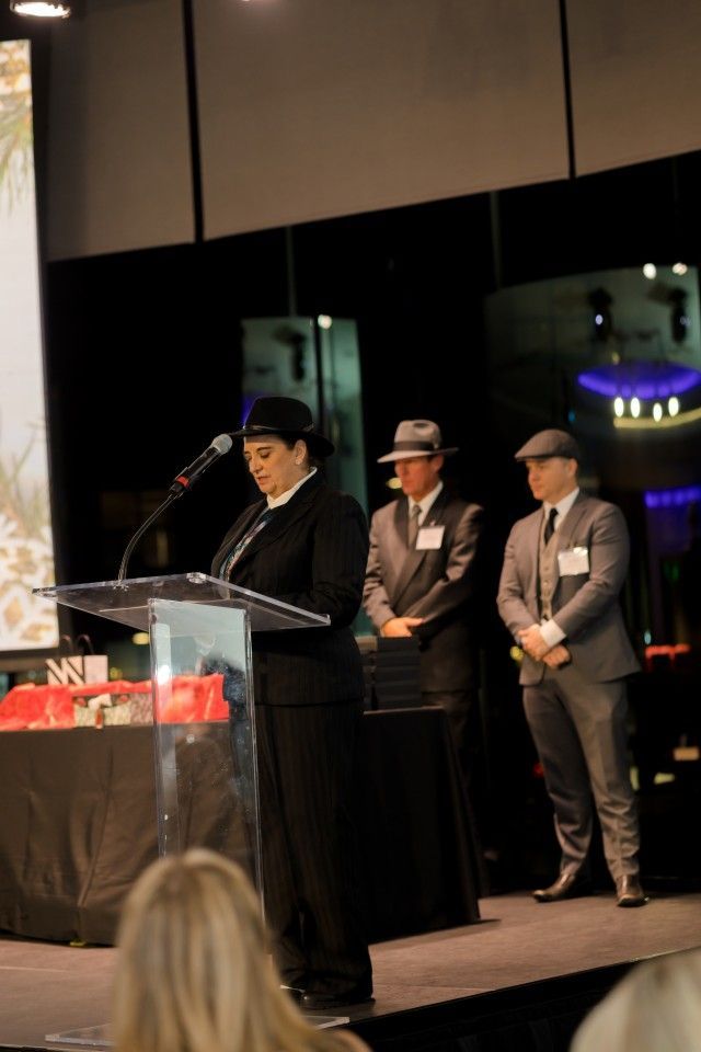Woman in suit speaking at podium, two men in fedoras and suits stand behind her on stage.