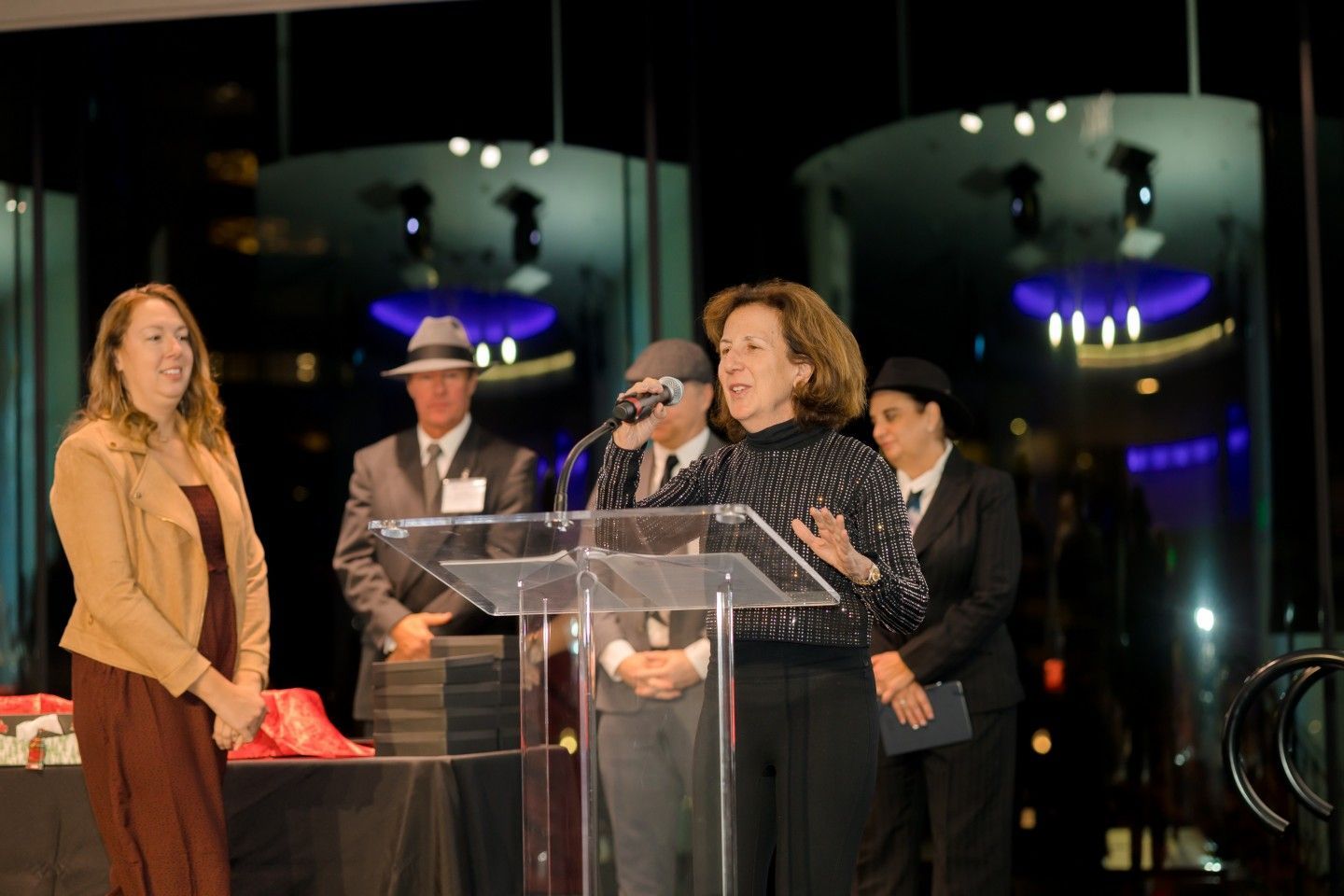 Woman speaking at podium, two women and two men behind her in a formal setting with city lights visible.