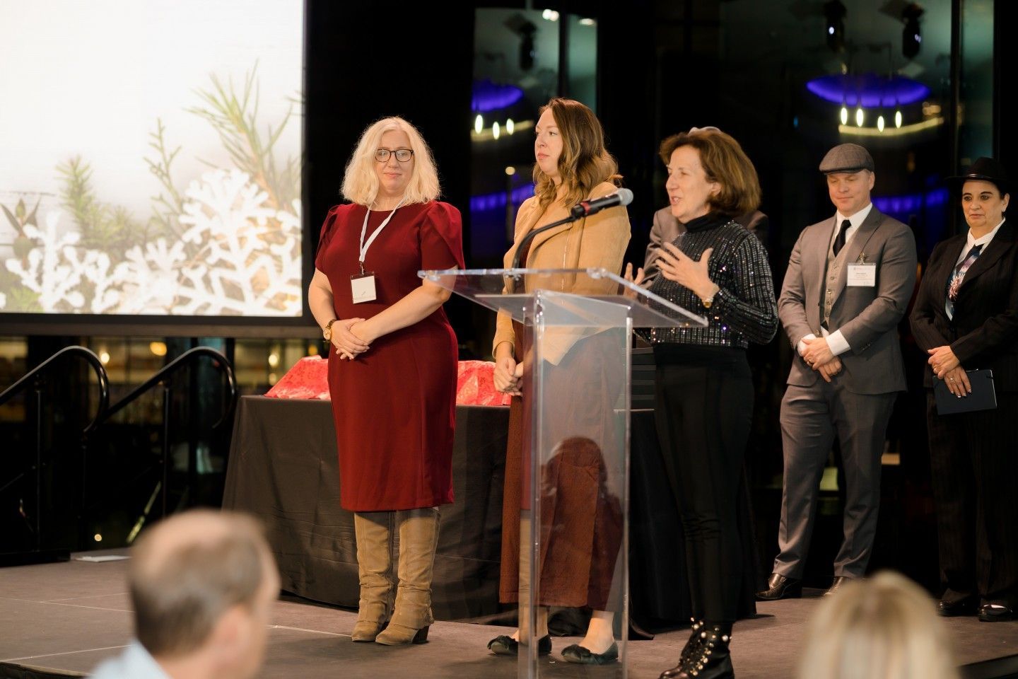 Three women and two men on stage with a podium and a screen. One woman is speaking.