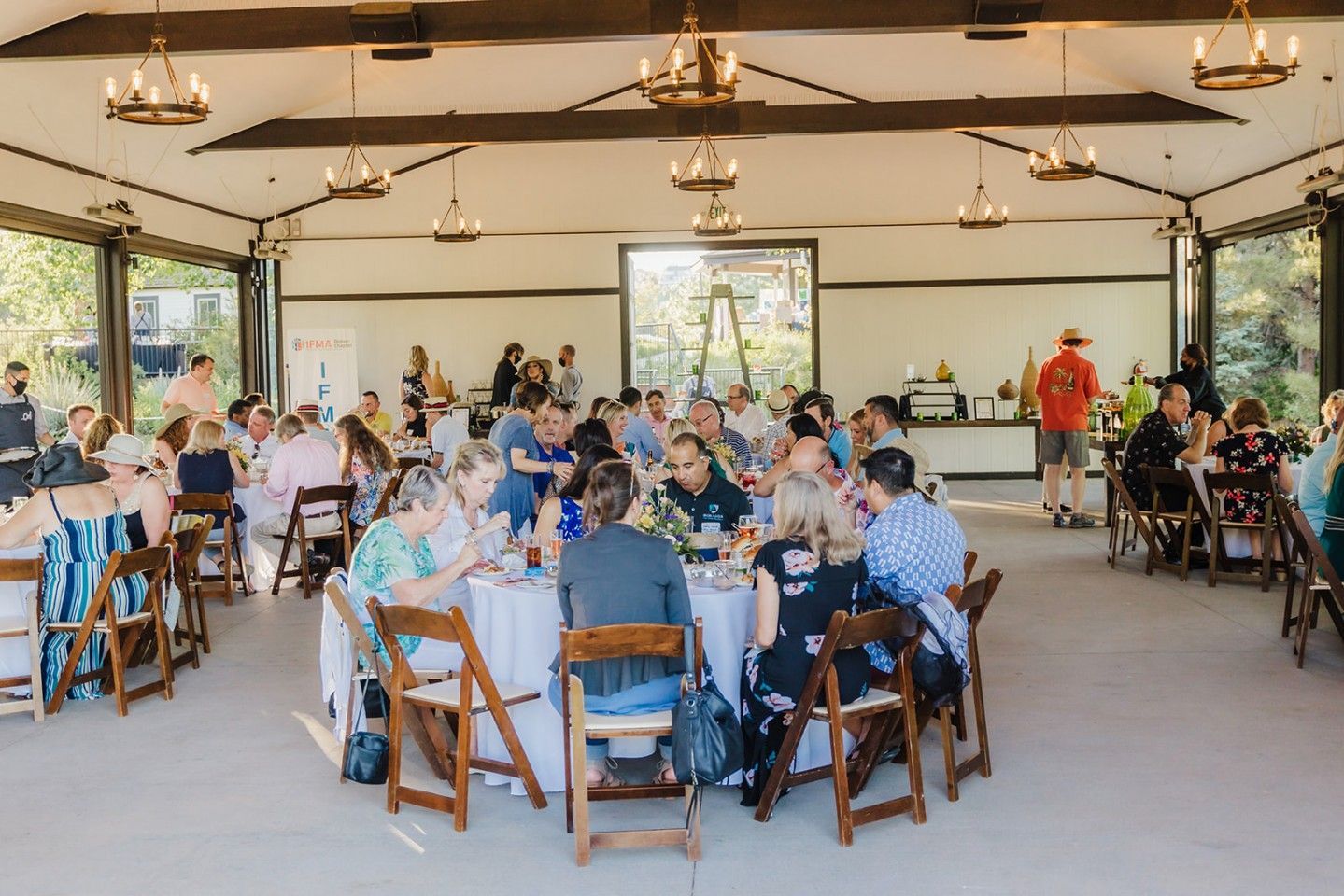 Large outdoor event with guests seated at round tables under a covered pavilion.