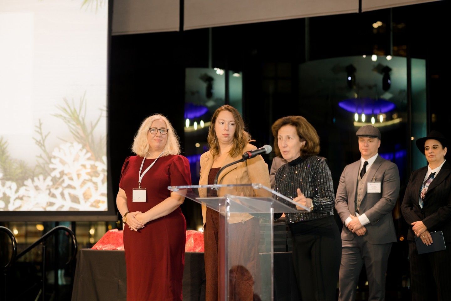 Five people stand at a podium, speaking at an event with a screen and decorations.