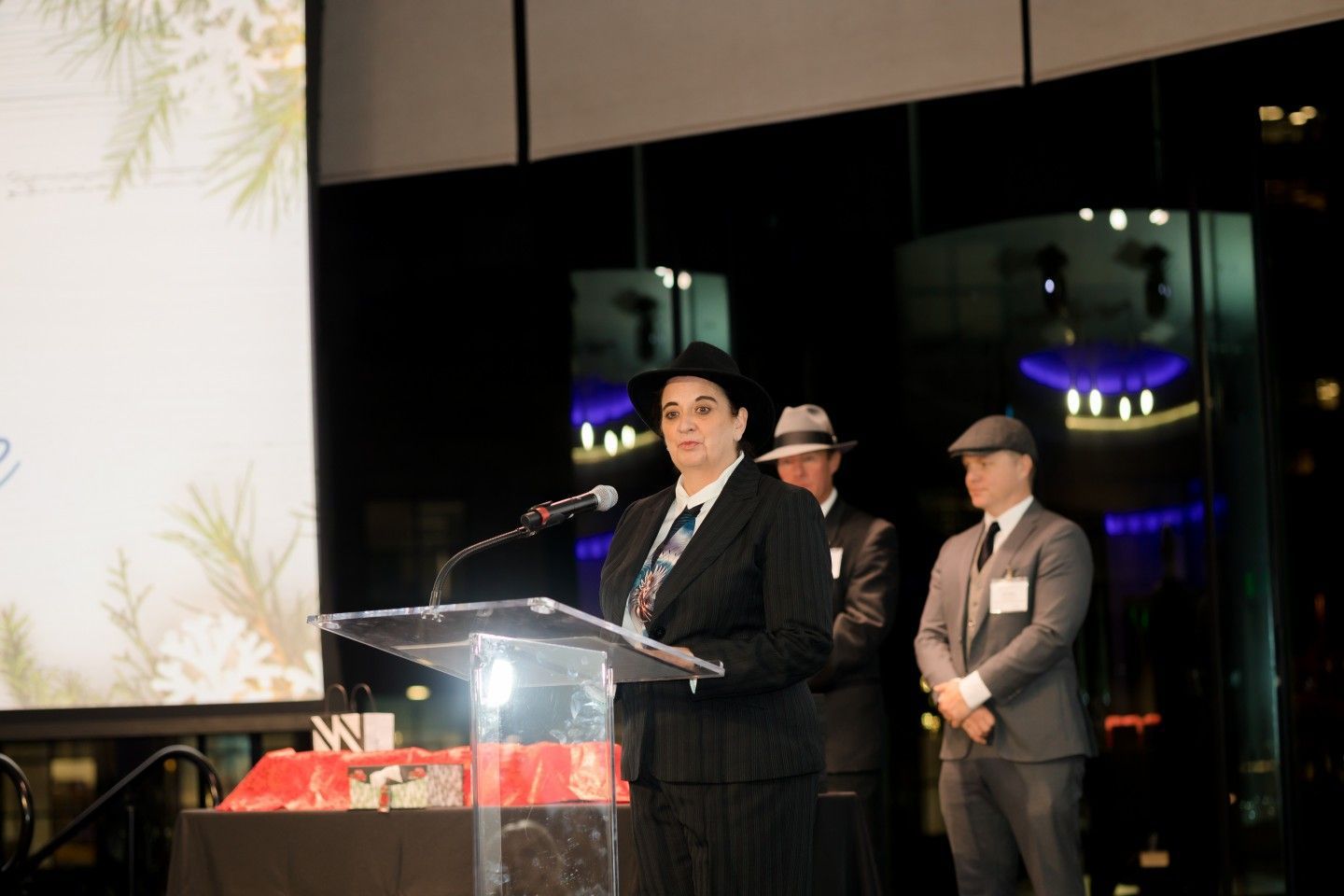 Woman in suit and hat speaks at a podium. Two men in suits and hats stand behind her. Nighttime event.