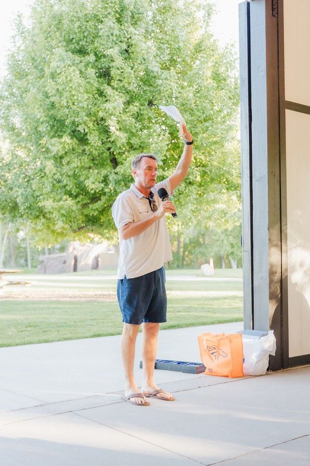 Man holding paper and microphone, gesturing on a patio. Overlooking lawn and trees, sunny day.