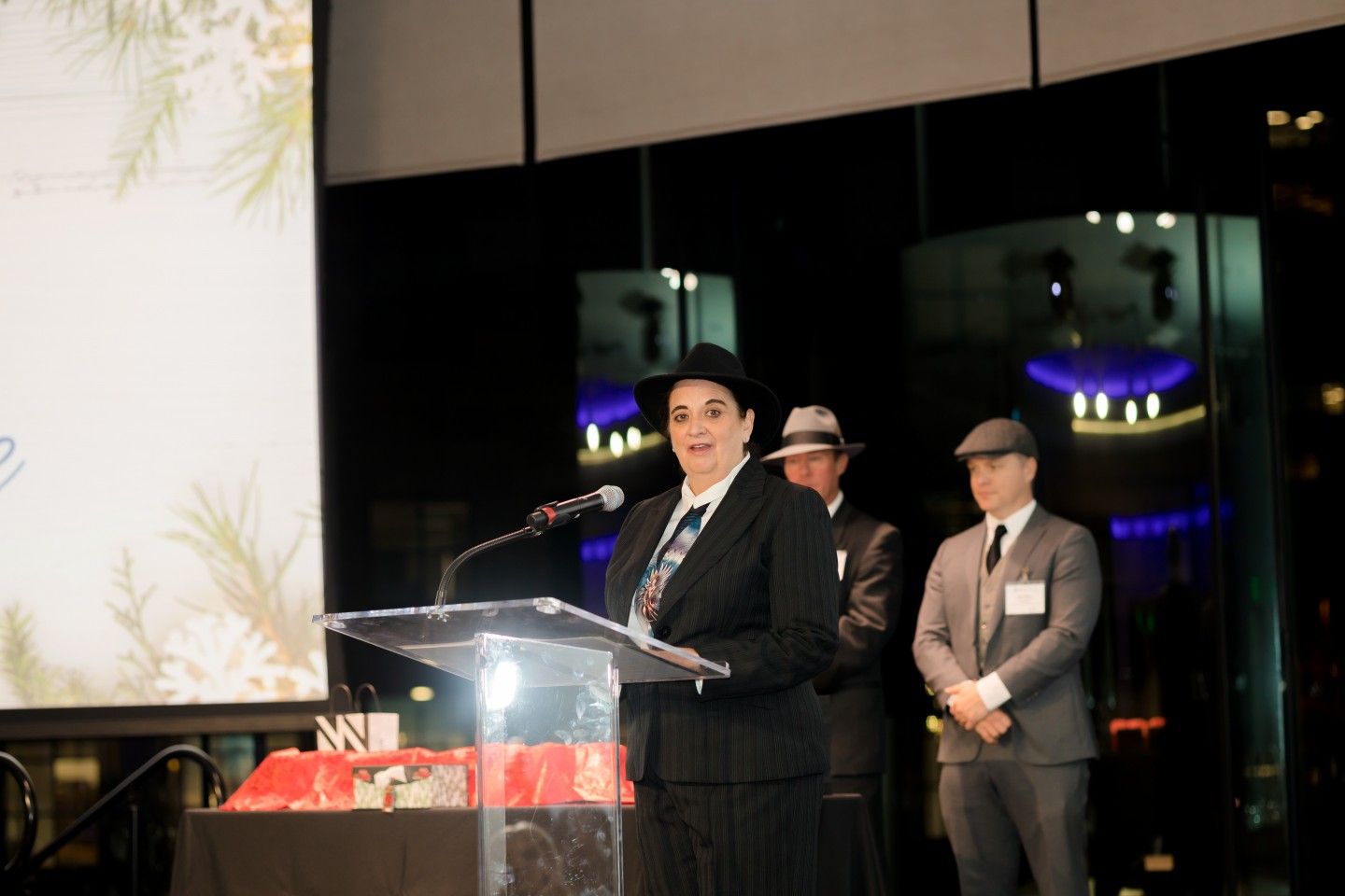 Person in a suit and hat speaking at a podium, two men in similar attire stand behind. Nighttime event.