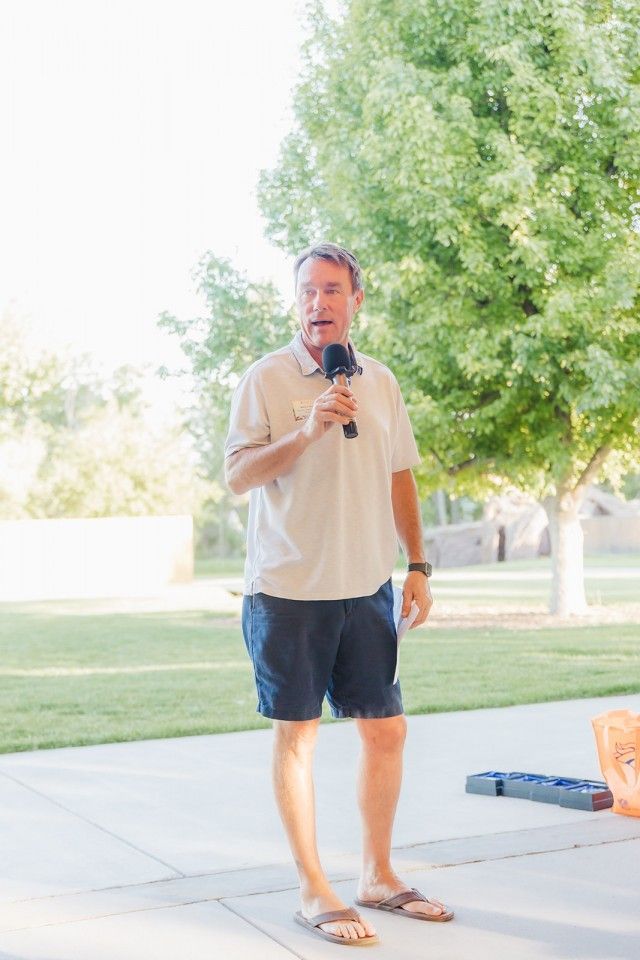 Man in shorts and sandals speaks into a microphone outdoors, near a tree.