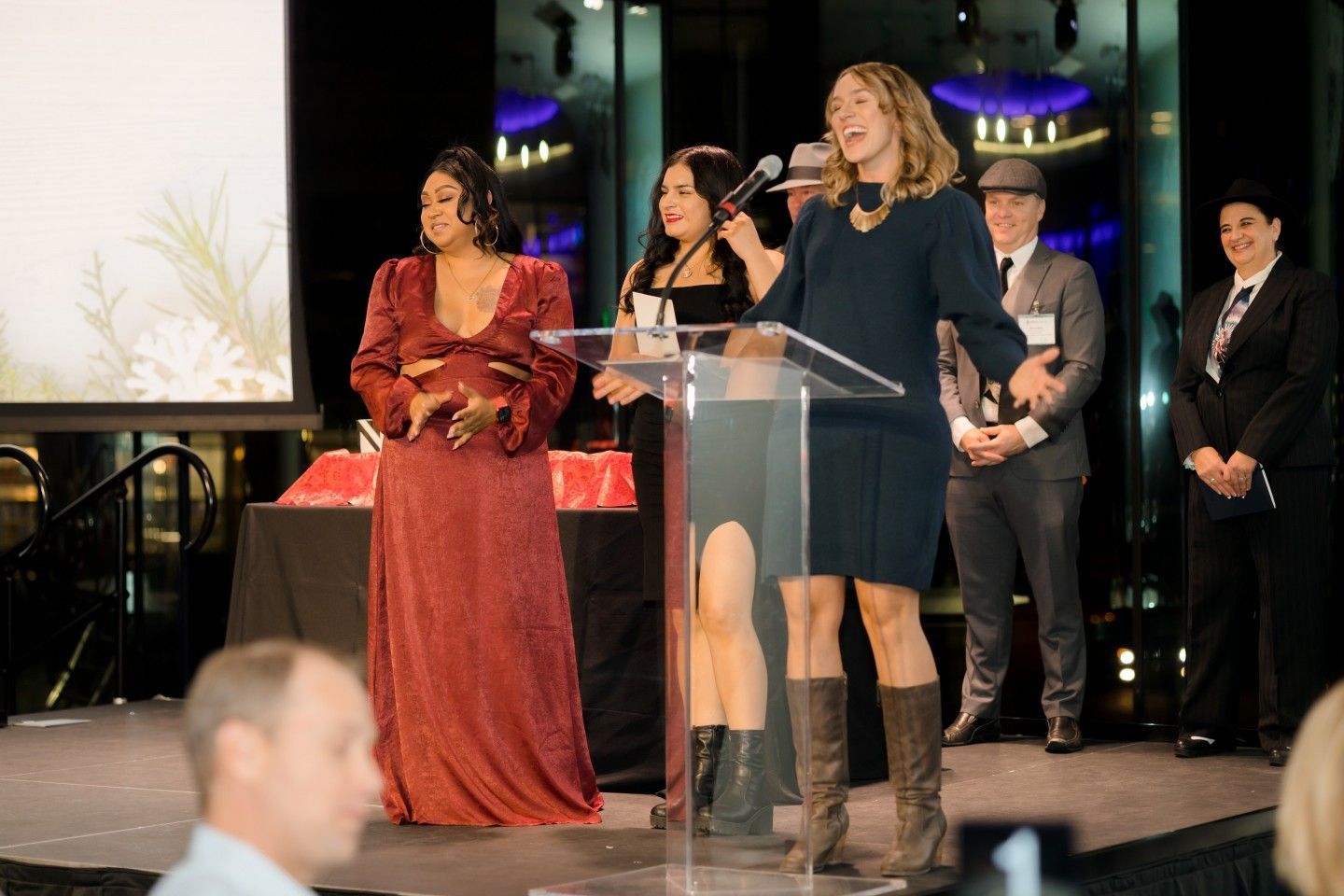 Woman speaking at podium, two women beside her, other people in background at event.