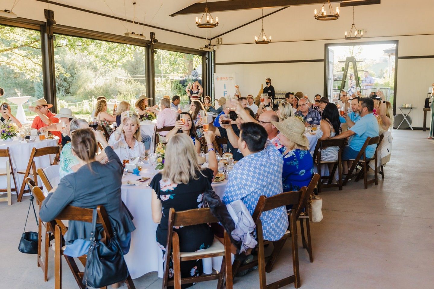Guests seated at tables inside a building with large windows. People are smiling and gesturing, a social gathering.