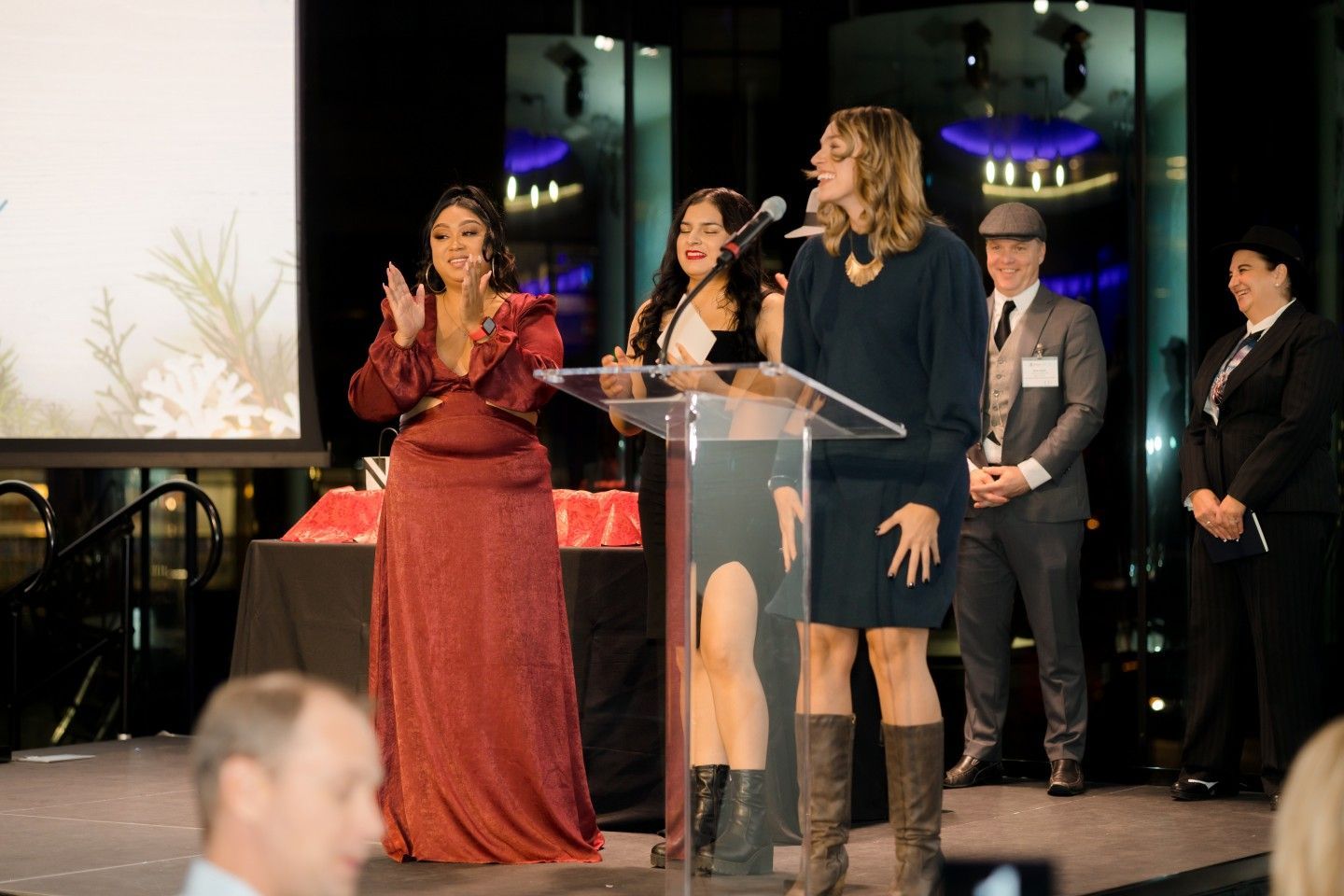 People on stage at event: woman speaks at podium, two clap, others watch. Indoors, festive setting.
