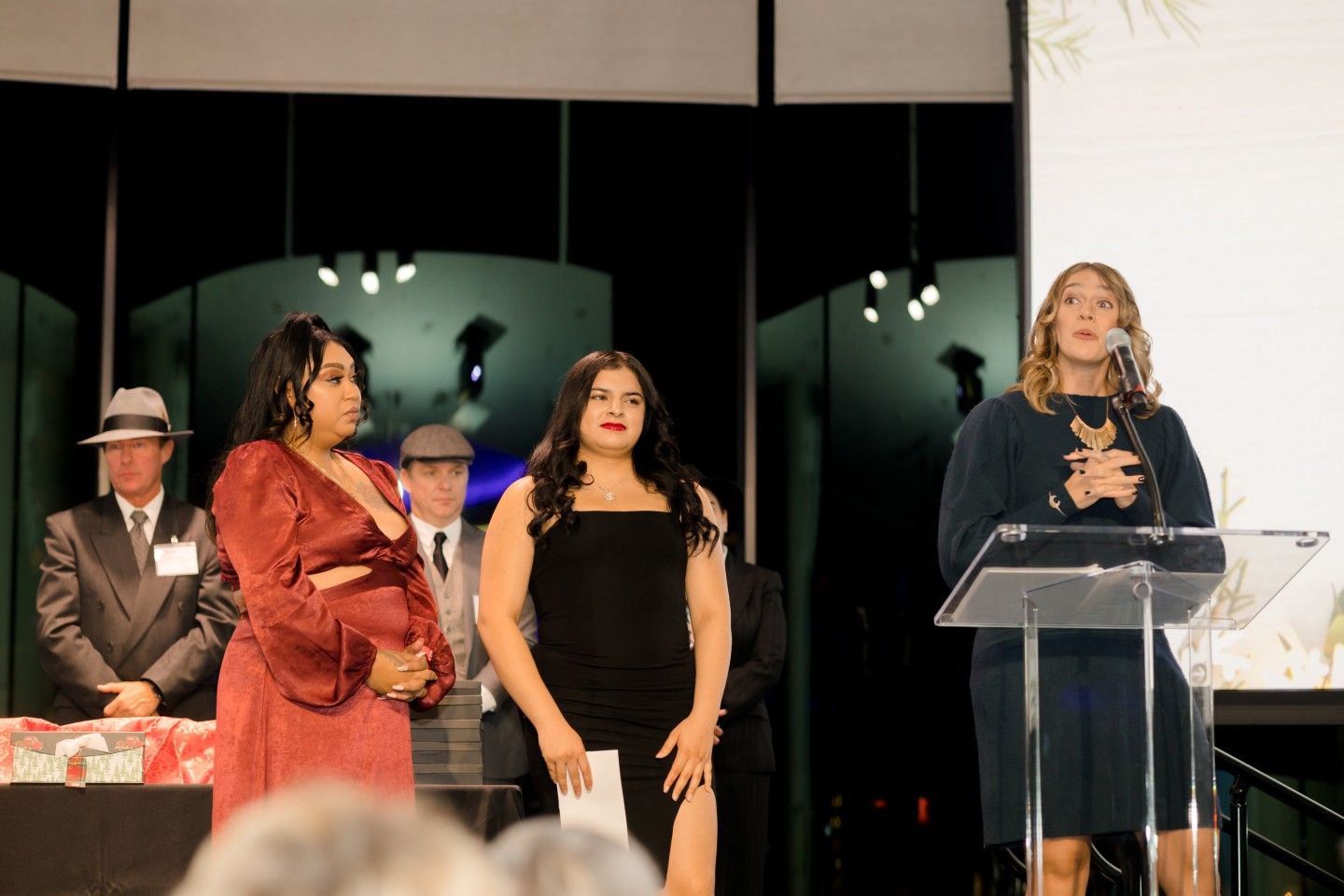 Three women standing at a podium on a stage with a man in the background. One is speaking, two are standing beside.