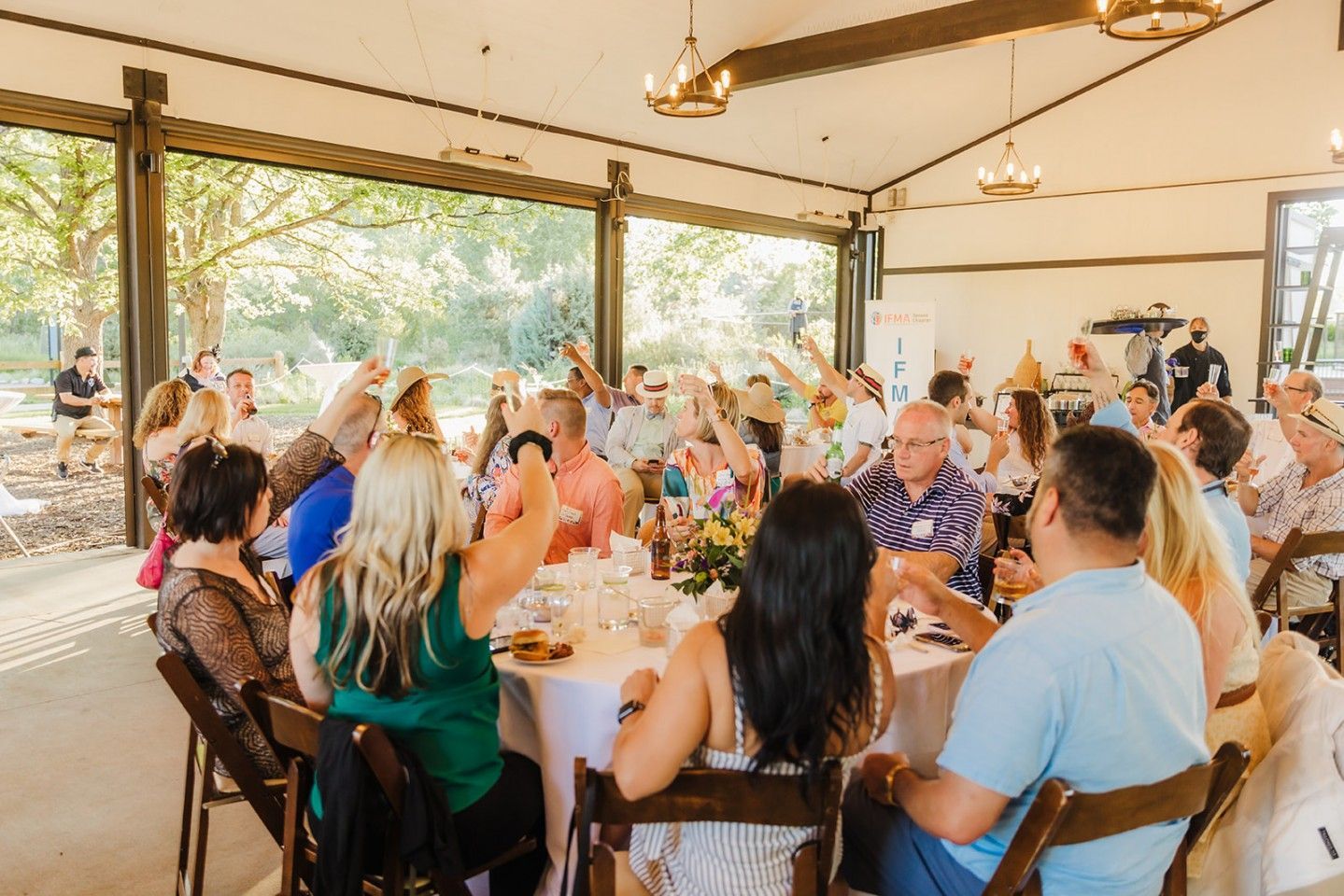 People gathered at tables in a bright room, raising hands and celebrating.