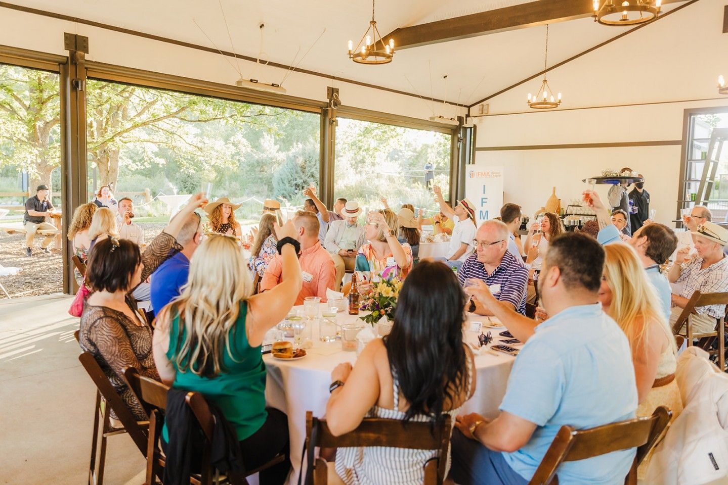 People at a celebratory gathering, raising glasses around a round table in an outdoor event space.