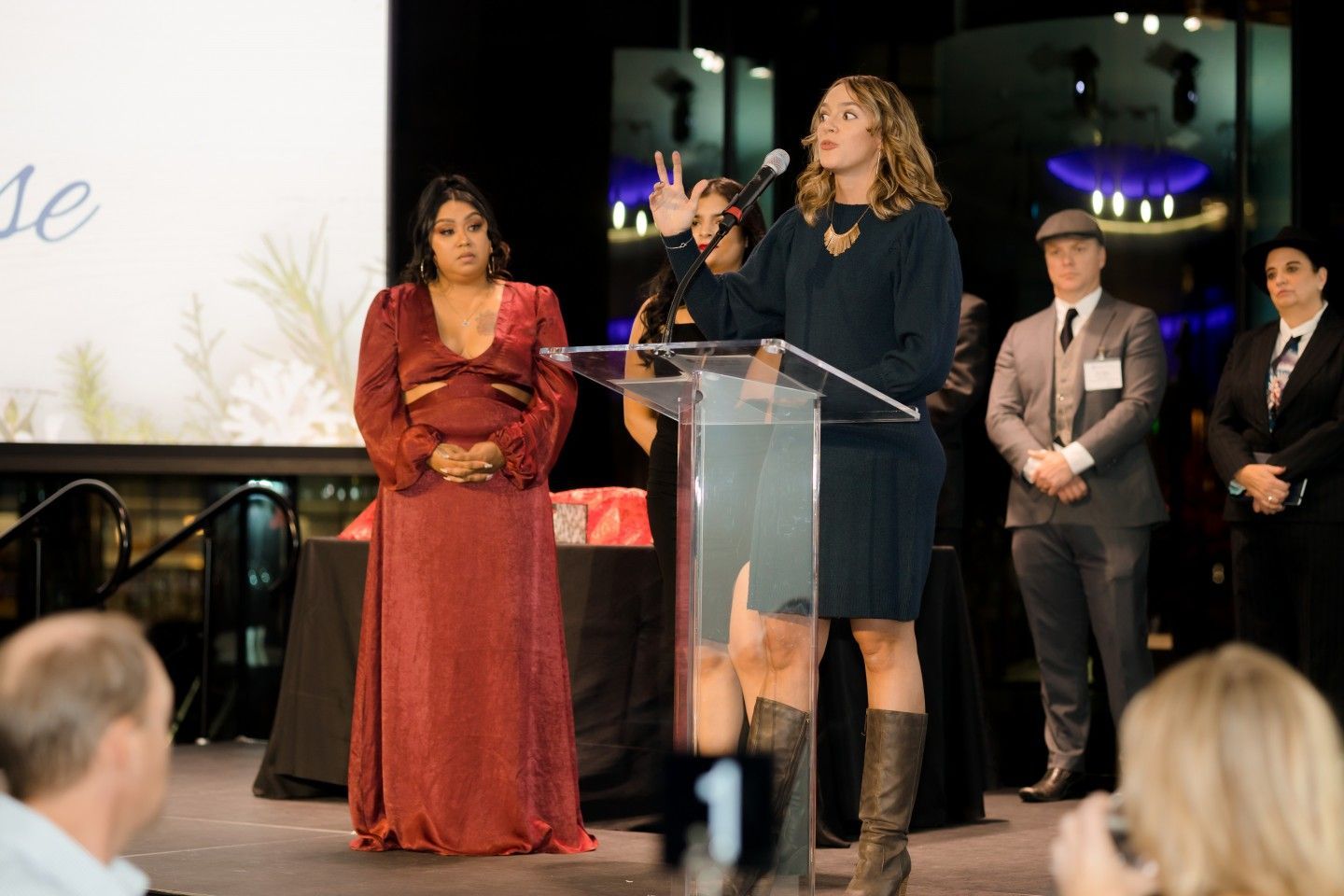 Woman speaking at podium, another woman stands to the side. Event setting, people in attendance.