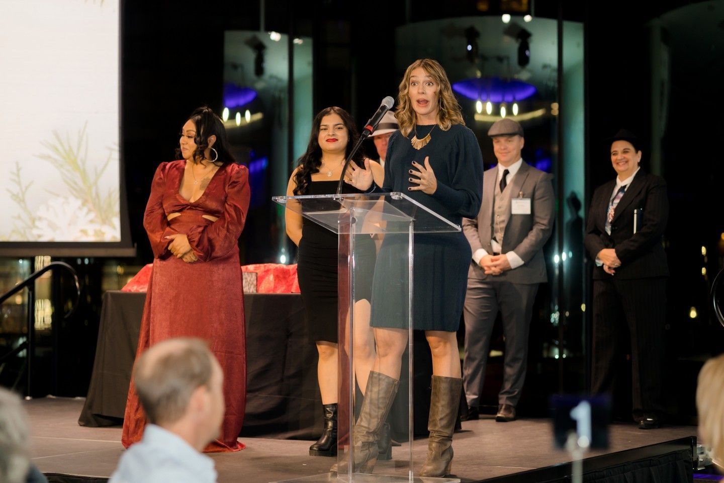 Woman speaking at a podium with others behind her at an event. Dark setting with stage, people, and awards.
