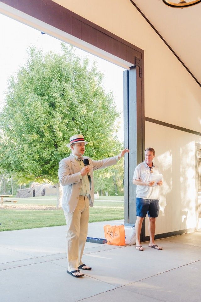 Man in suit with microphone gesturing outdoors; another man standing beside him.