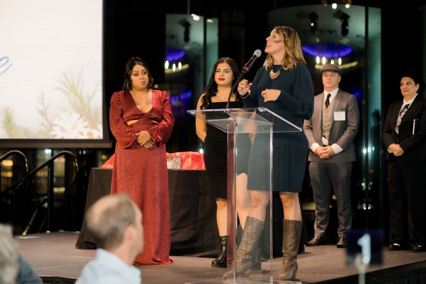 Woman speaks at a podium; two women and two men stand behind her. Stage setting, various outfits.