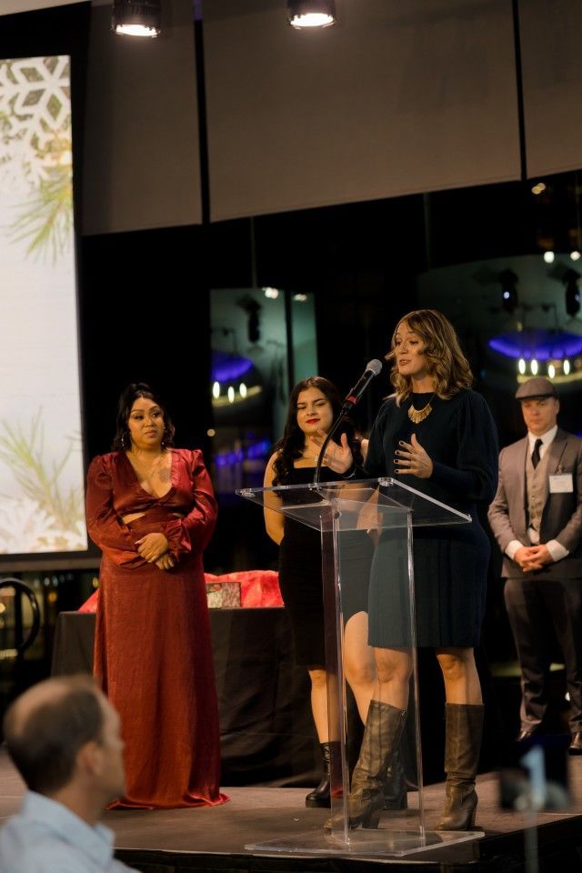 Woman speaking at podium, two women standing nearby. Formal event, holiday decorations, dark clothing.