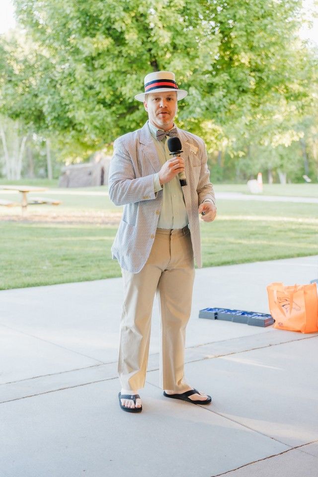 Man in seersucker suit, hat, and bow tie speaks into a microphone outdoors.