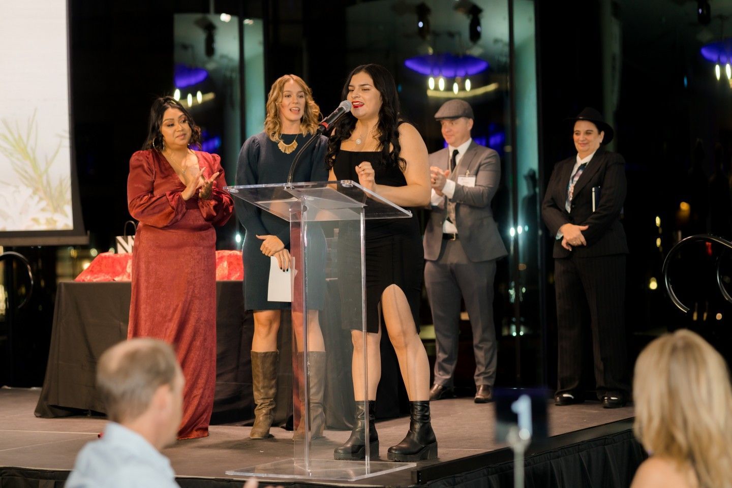 Woman in black dress speaks at a podium. Others clap and watch on a stage with a city view.