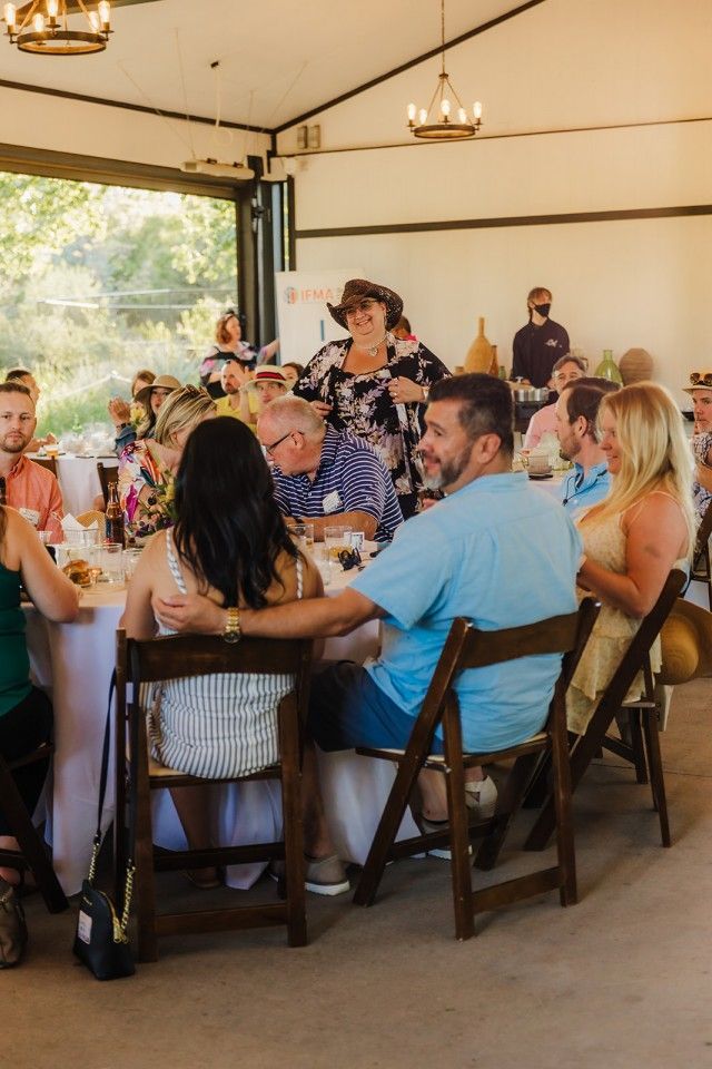 People seated at tables inside a building with large windows, some looking at a woman speaking.