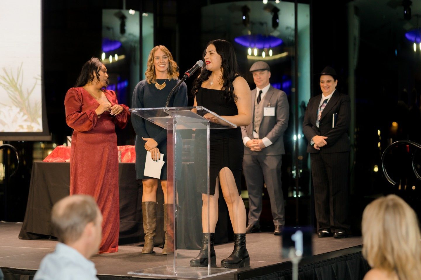 Woman speaking at podium during awards ceremony with others on stage.