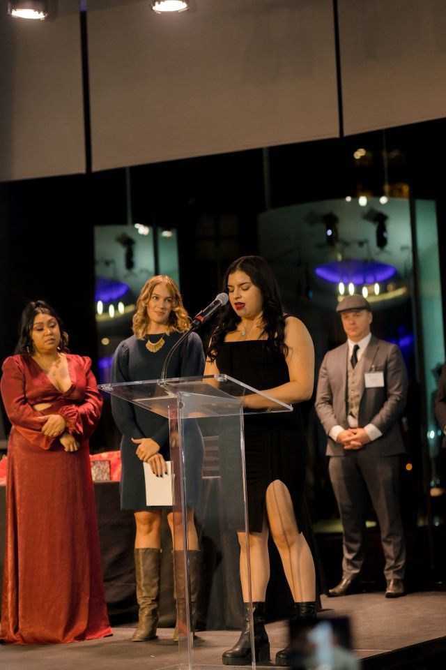 Four people on stage at an event: a woman speaking at a podium, others standing by.