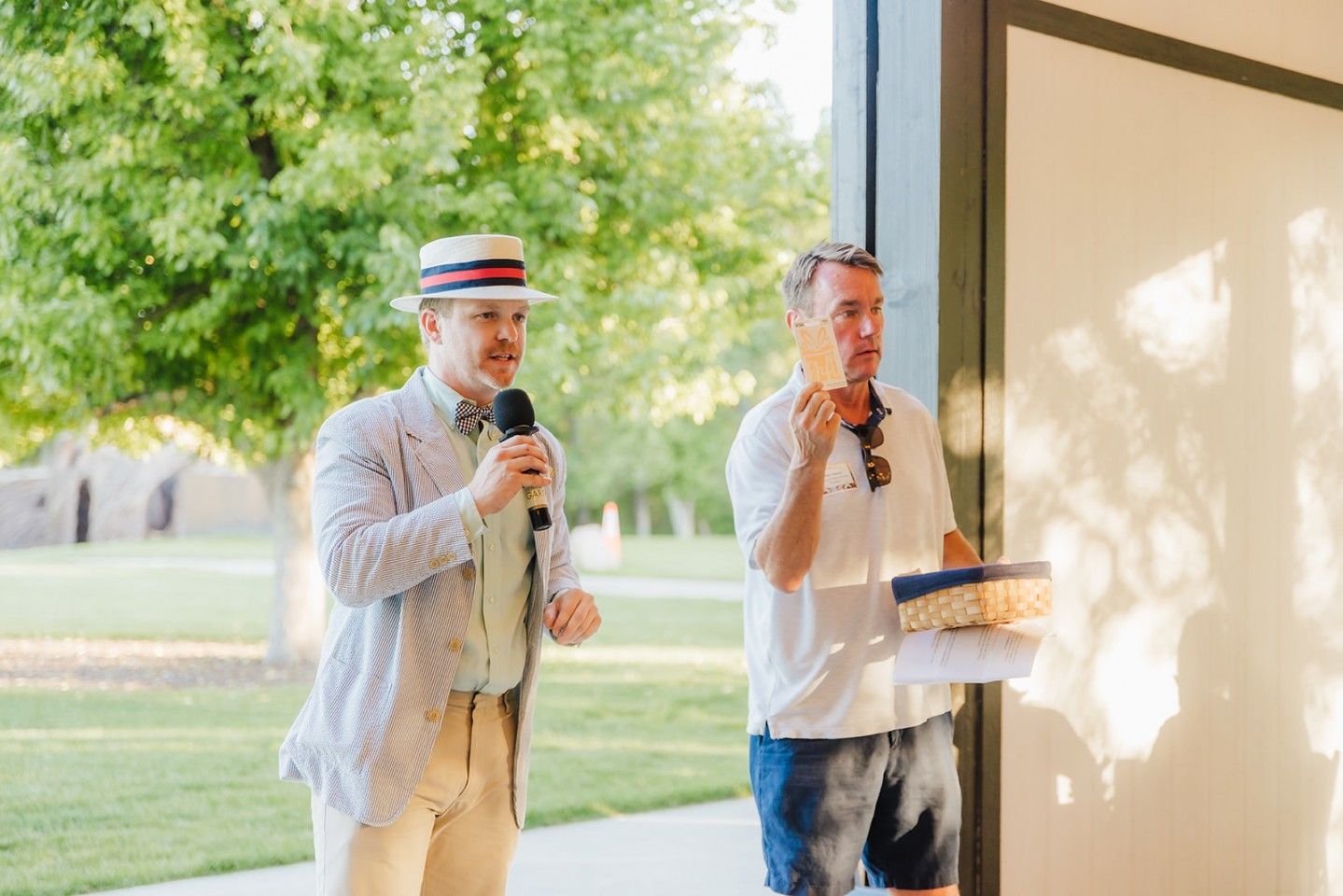 Two men outdoors, one in a hat speaking into a microphone, the other holding food.