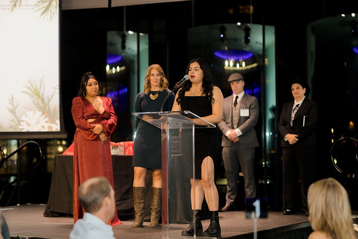 Woman speaking at podium during awards event; four people standing, city backdrop.