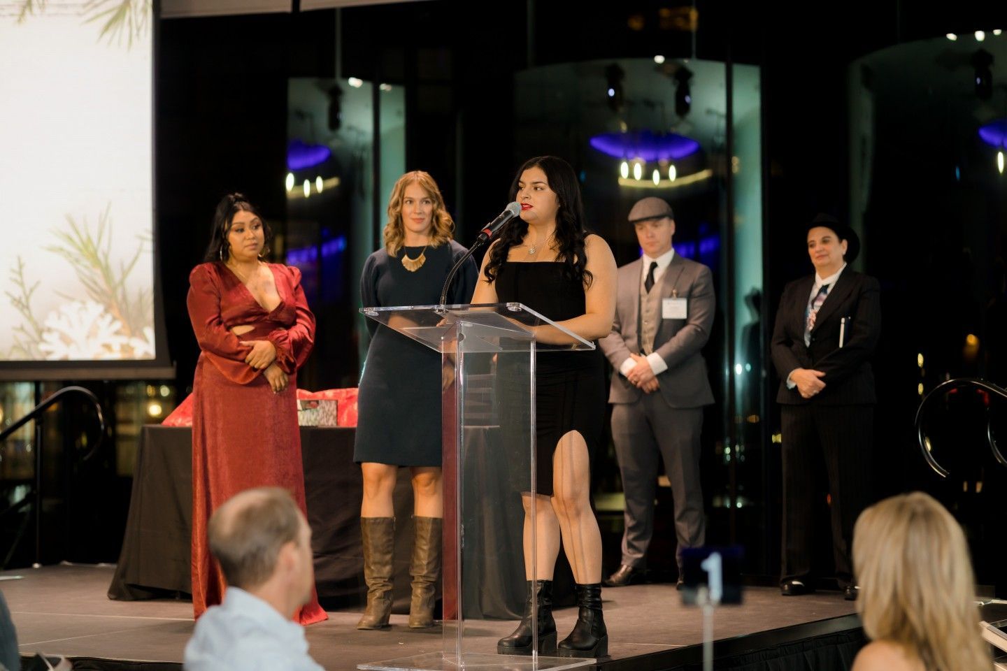 People on stage at an awards event. Woman speaks at podium, others stand nearby, watching.