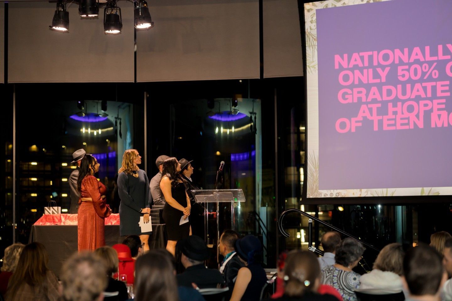 People on stage at an event, speaking with a screen showing text. Audience in foreground. Dark setting with city view.