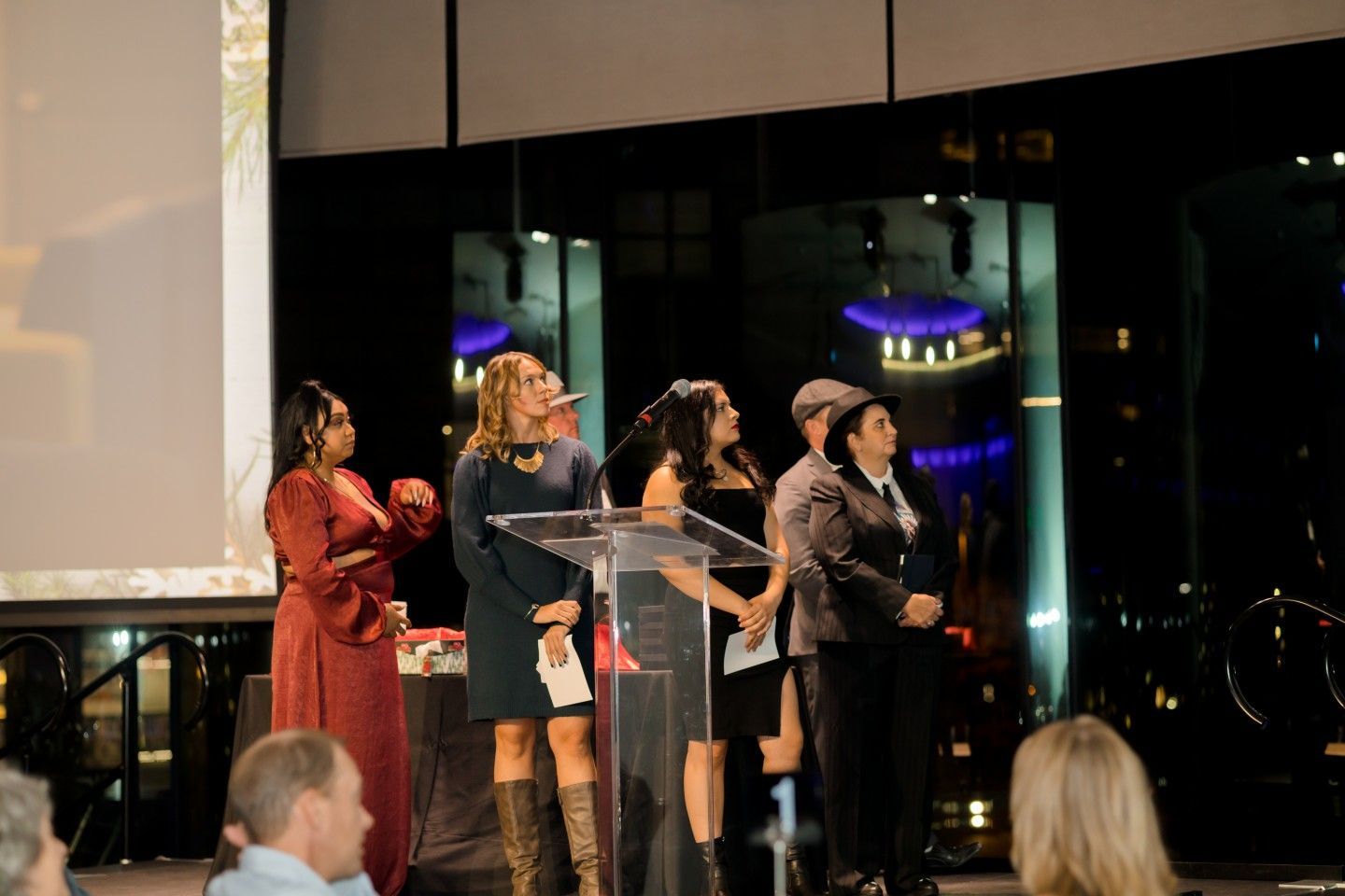 People stand at a podium, looking up. A large screen and city lights are visible in the background.