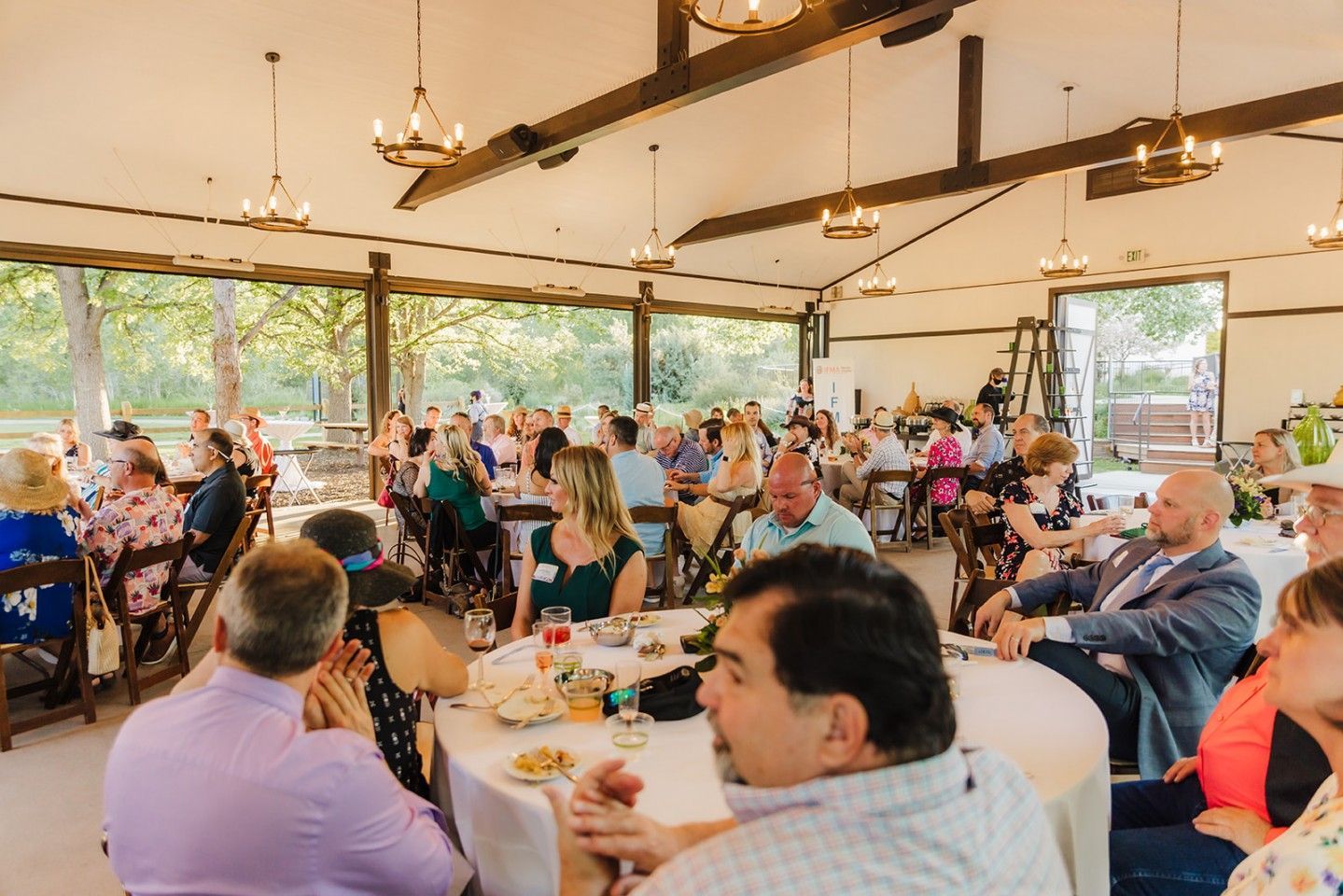 Large group of people seated at round tables in a pavilion. Many are listening while others eat and talk.
