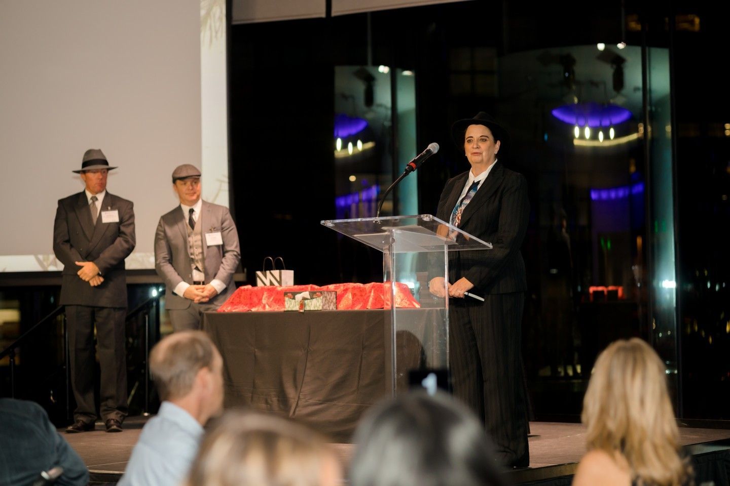 Woman in a suit speaks at a podium; two men in suits and hats stand behind her. Awards on a table.