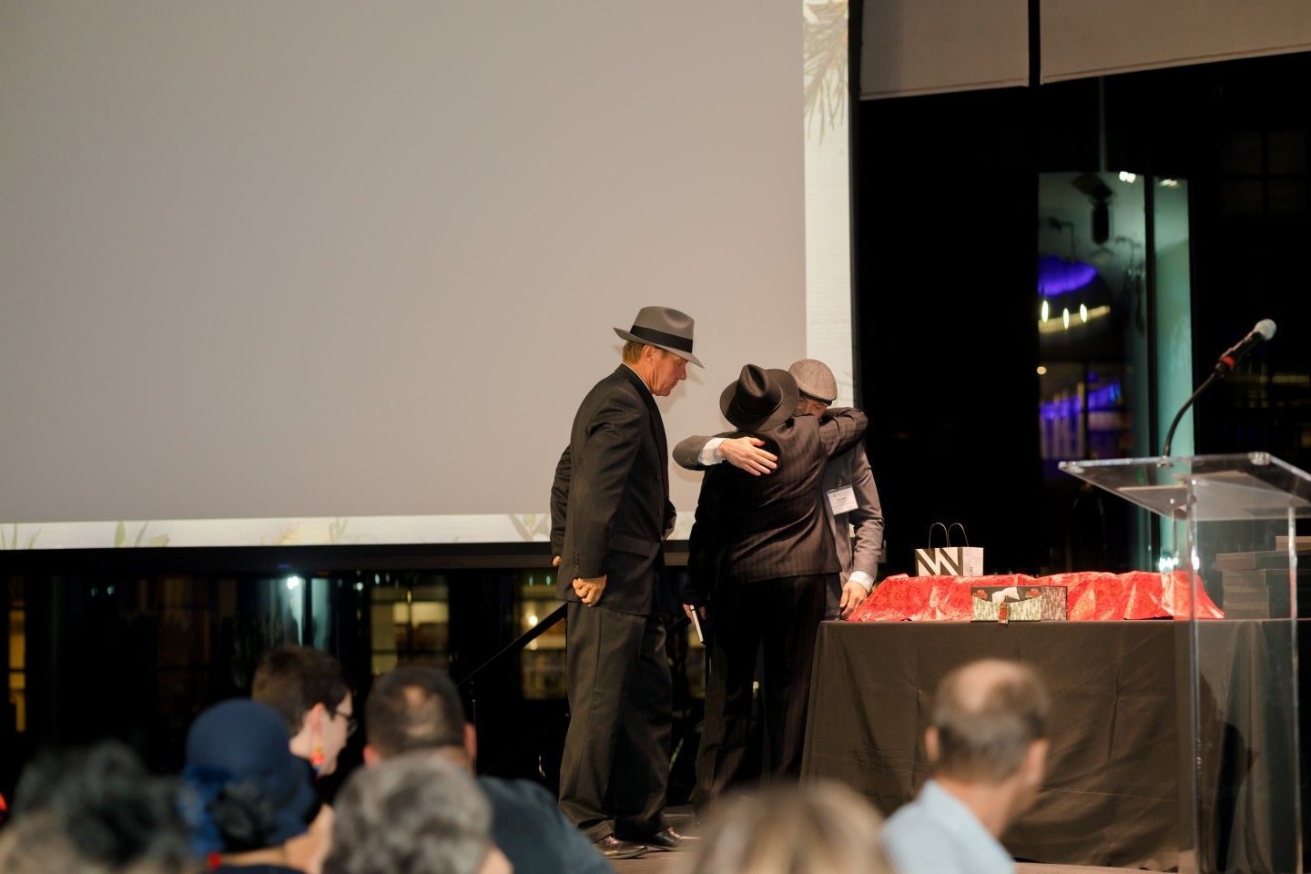 Man in suit hugs another on stage with trophies, next to a podium, dark setting.
