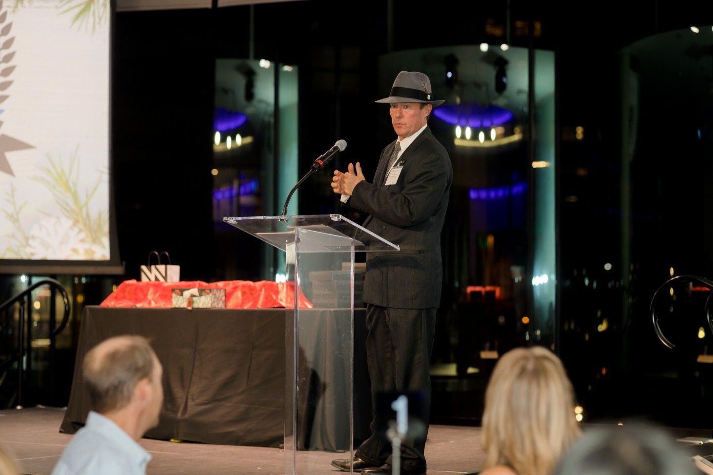 Man in suit and fedora at podium, speaking at event. Red table cloth, dark background.