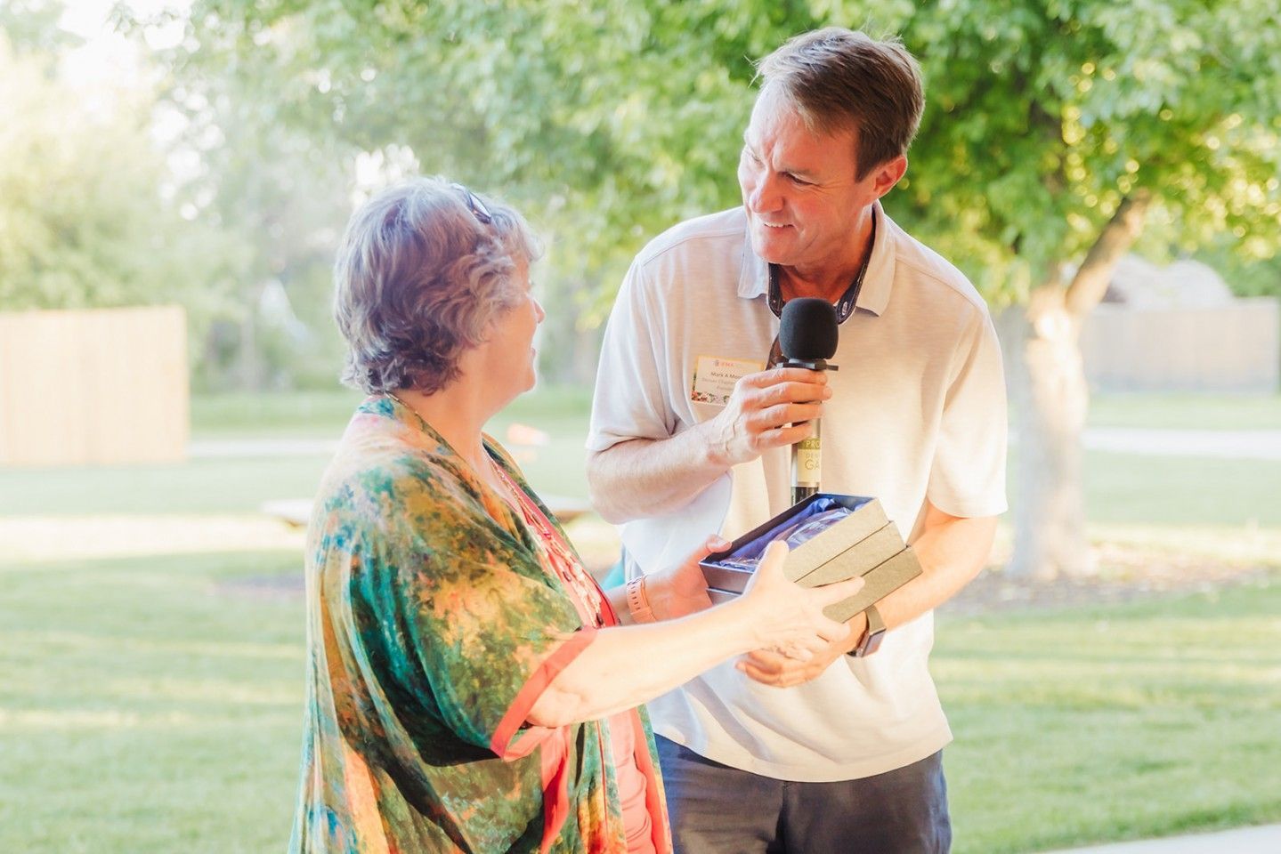 Man handing a gift to a woman, holding a microphone outdoors.