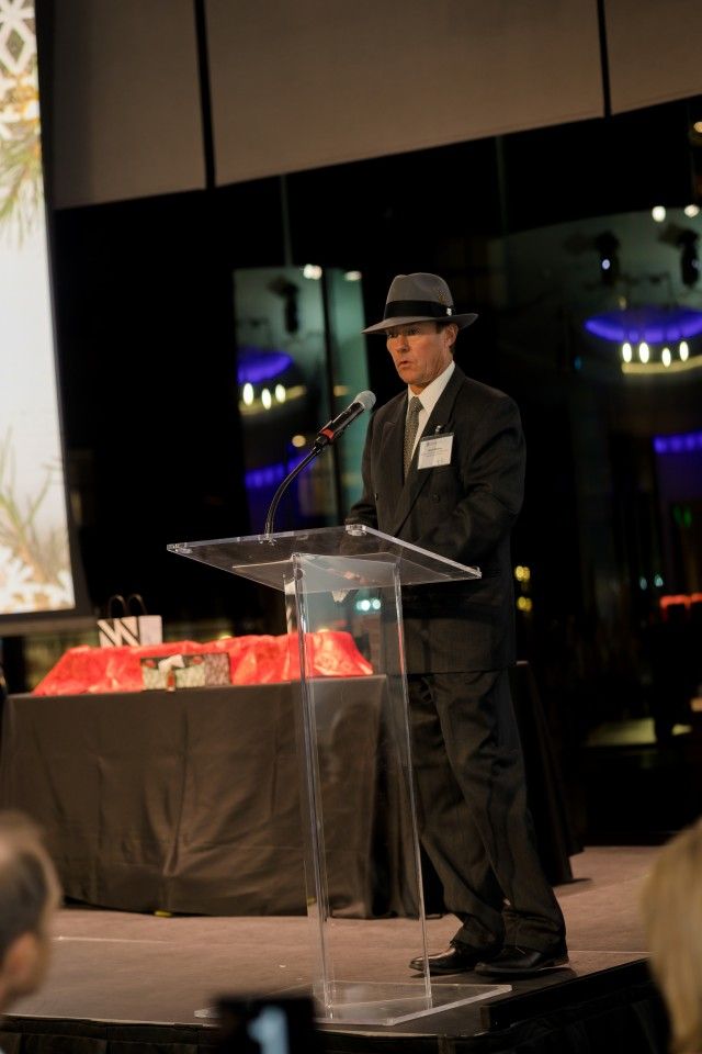 Man in suit and hat speaking at a podium, stage setting.