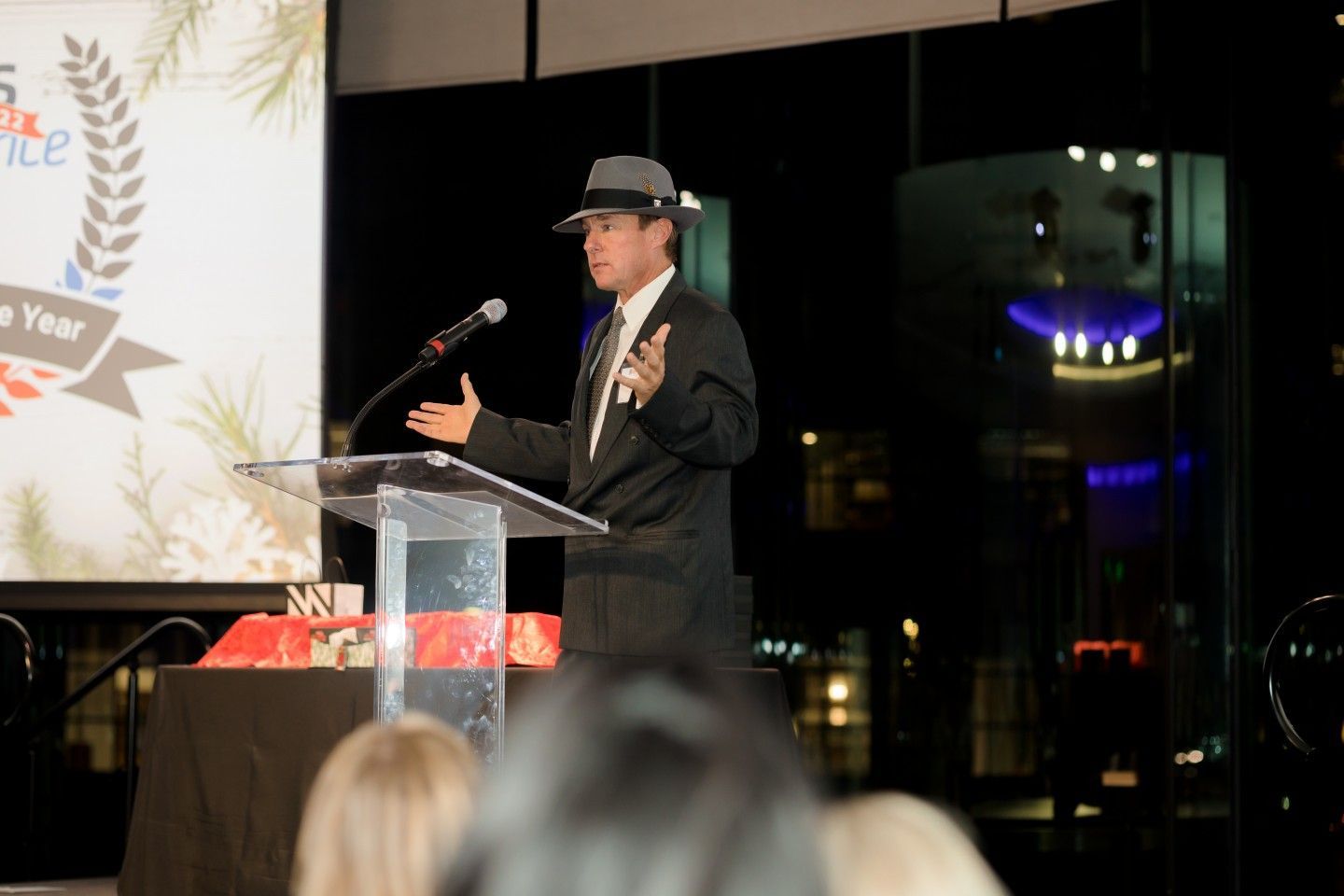 Man in a suit and fedora speaks at a podium with a banner behind him.