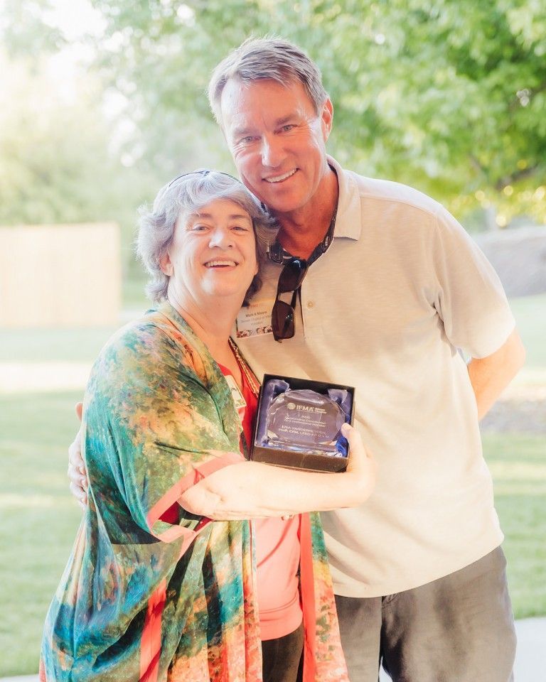 Woman holding plaque, embraced by man outdoors, both smiling.