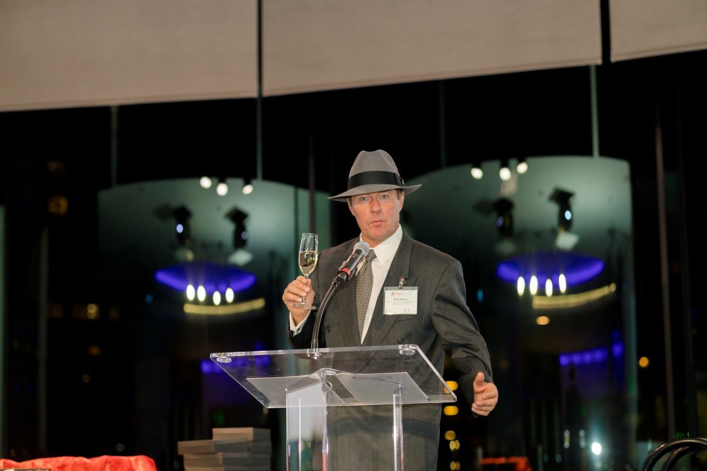 Man in a suit and fedora holding champagne, speaking at a podium in a formal setting.