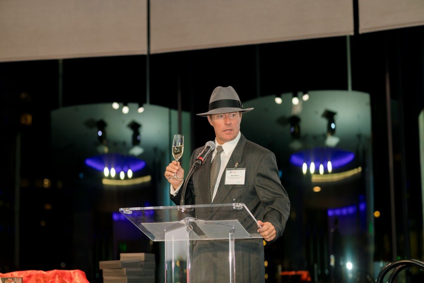 Man in suit and fedora raises champagne glass at podium, lit interior, night view in background.