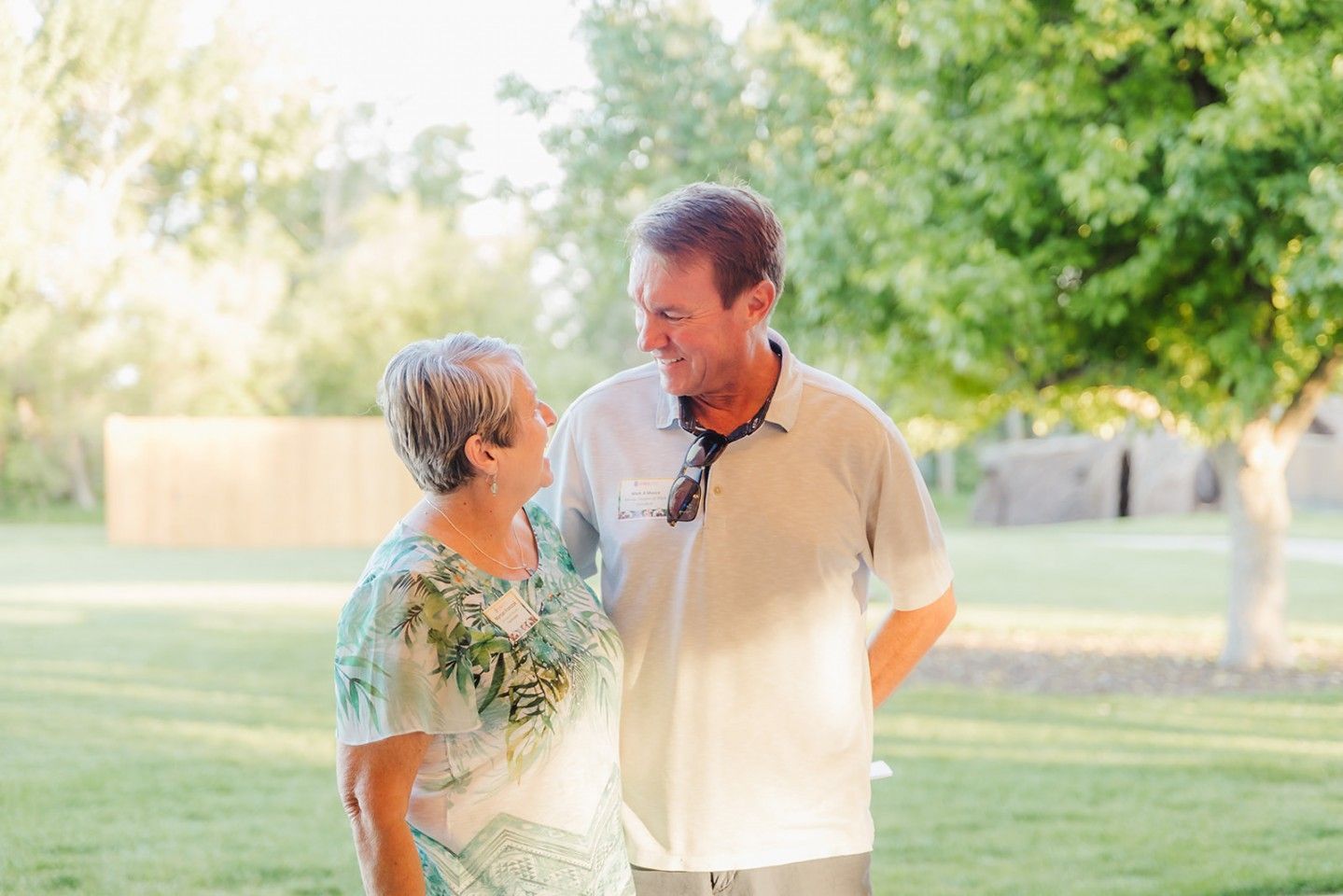 Woman in floral top and man in polo shirt smiling at each other outdoors, next to a tree.