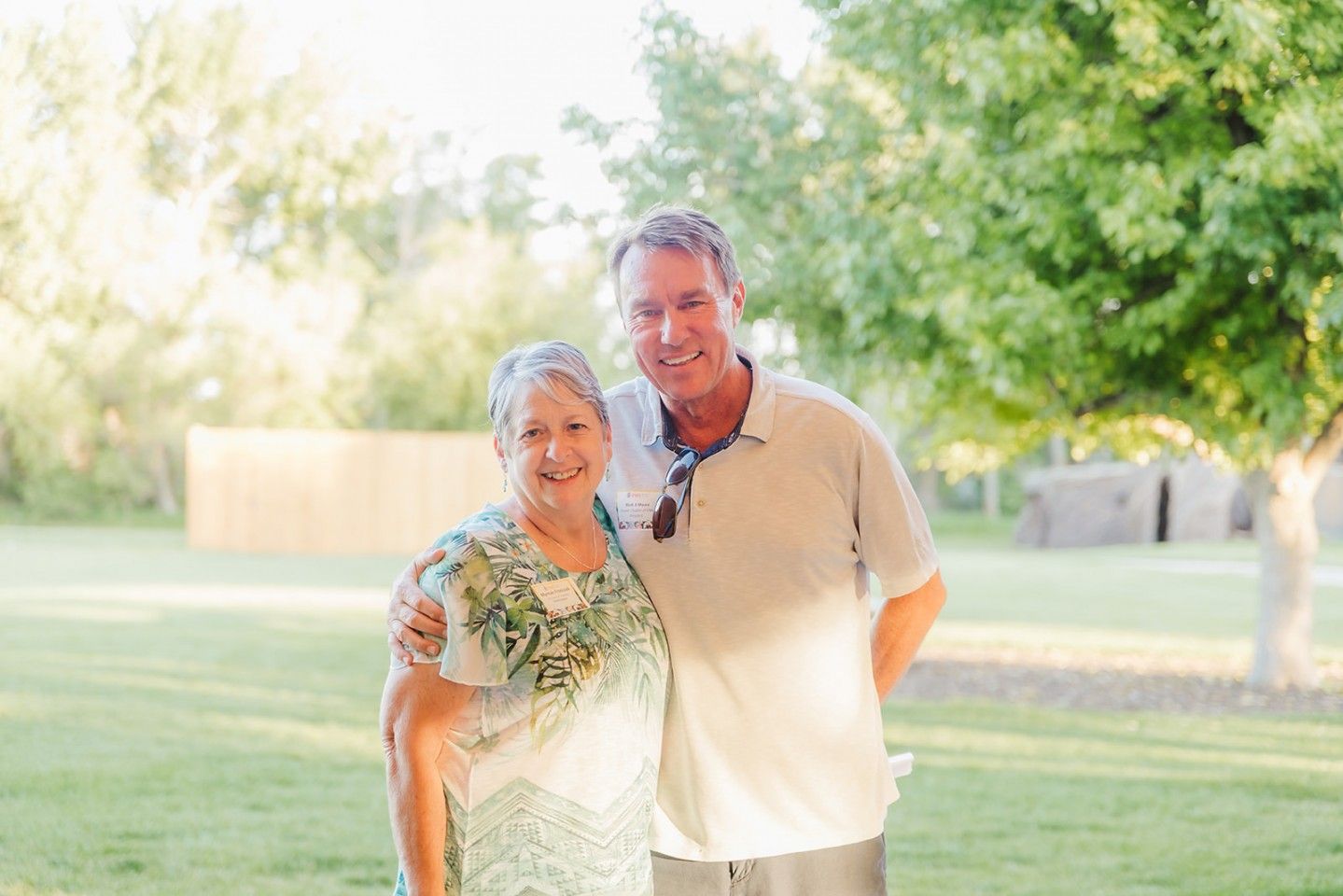 Woman and man with arms around each other, smiling outdoors. Green dress, tan shirt, trees and grass in background.