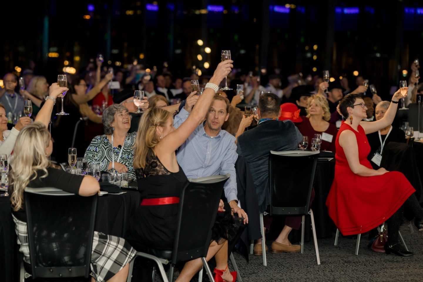 People toasting with champagne glasses at a formal event, some raising arms. Dark setting with city lights in background.