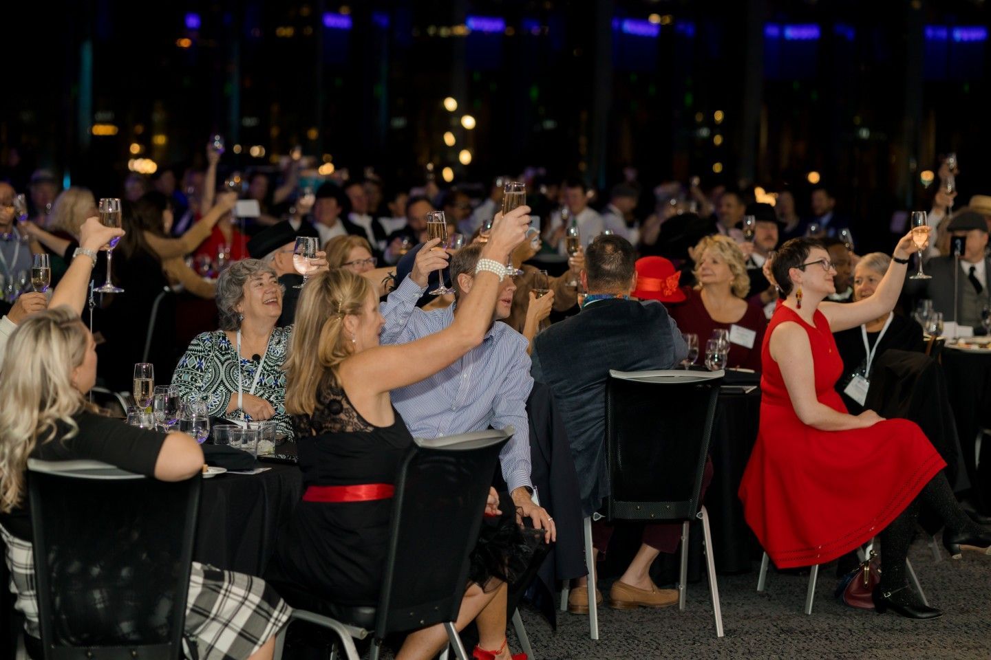 People at a banquet raising champagne glasses in a toast, dark room with city lights in background.