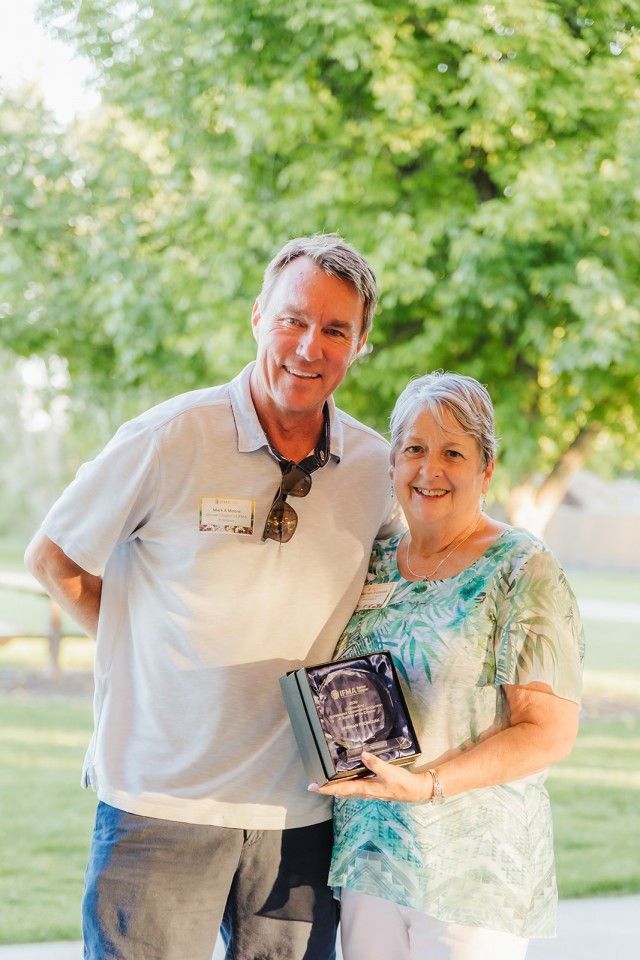 Man and woman smiling, holding an award. Outdoors, green foliage in the background.