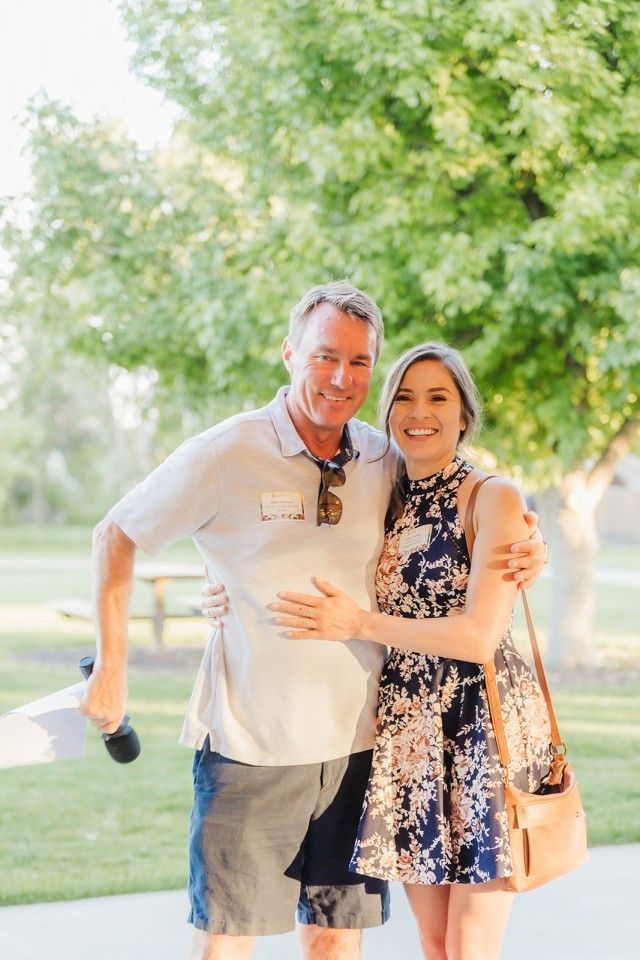 A man and woman standing together outdoors smiling. Man holds microphone. Woman has her arm around the man.