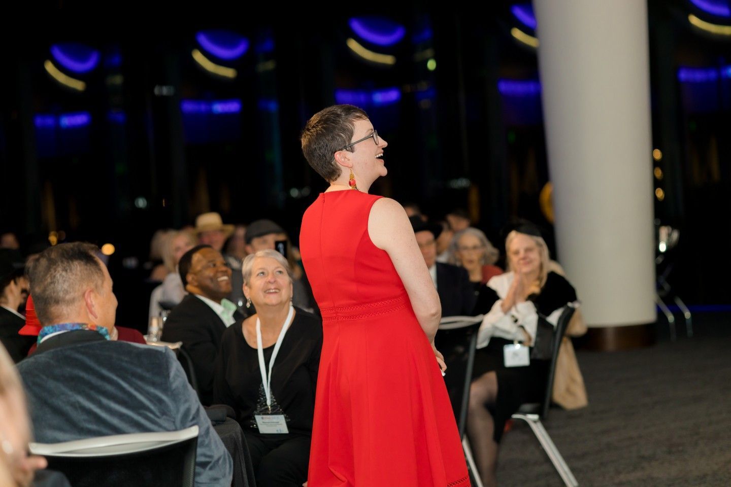Woman in a red dress speaking at an event; people in the audience listen and applaud.