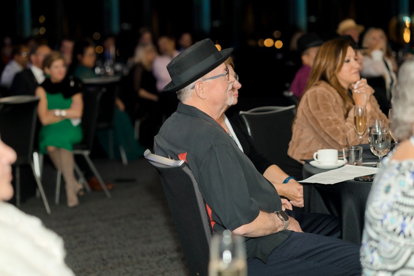 Man in black hat at a formal event, seated and looking to the side. People seated in background.